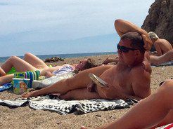 An elderly nudist sunbathes and reads a...